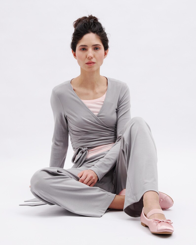 May include: A light gray wrap top with long sleeves and a matching pair of wide-leg trousers. The outfit is layered over a pale pink undershirt. The ensemble is paired with light pink ballet flats featuring a small bow and a strap across the top.