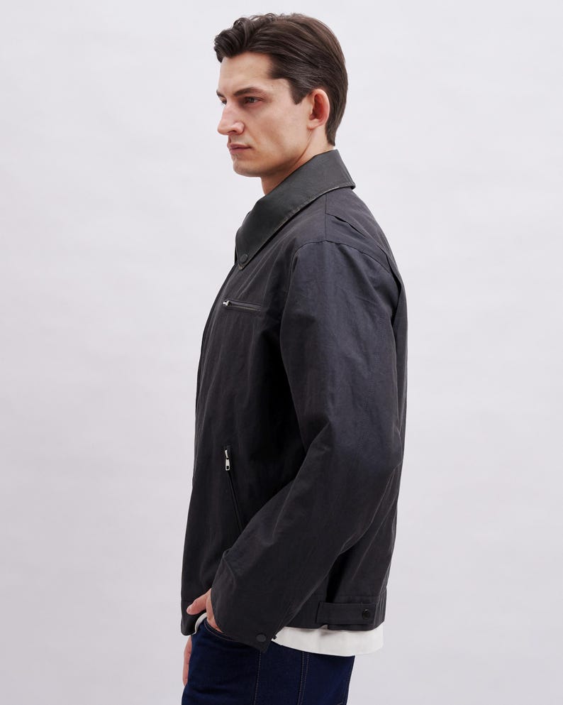 K&ouml;nnte beinhalten: Dunkelgraue Jacke mit Kragen und Rei&szlig;verschluss. Die Jacke hat einen dezenten Glanz und eine taillierte Passform. Das Model tr&auml;gt ein wei&szlig;es Hemd darunter. Die Jacke scheint aus strapazierf&auml;higem Material zu sein.