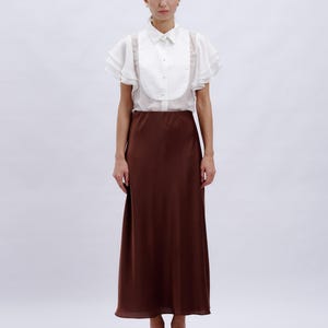 May include: A white blouse with ruffled sleeves and a brown midi skirt. The blouse has a high collar and a button-up front.