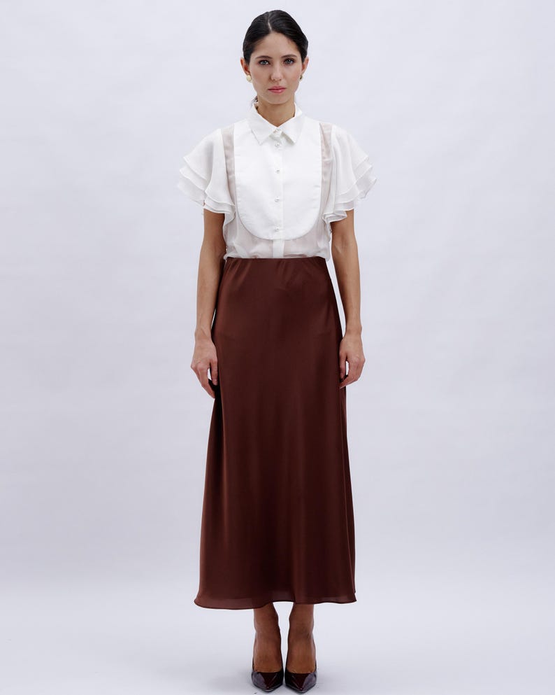 May include: A white blouse with ruffled sleeves and a brown midi skirt. The blouse has a high collar and a button-up front.