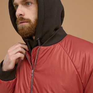 May include: A red bomber jacket featuring a dark brown hood and matching dark brown ribbed cuffs. The jacket has a full-length silver-toned metal zipper closure down the center front and raglan sleeves with a smooth, slightly reflective finish.