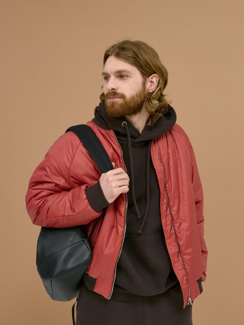 Peut inclure: Un blouson bomber rouge avec une fermeture &eacute;clair frontale port&eacute; sur un sweat &agrave; capuche marron fonc&eacute;. Un sac &agrave; dos noir est port&eacute; sur une &eacute;paule. La veste pr&eacute;sente des poignets c&ocirc;tel&eacute;s et une finition en tissu synth&eacute;tique lisse et brillant.