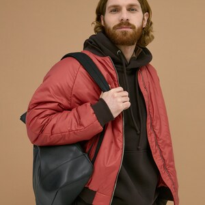 May include: A red bomber jacket with a front zipper closure and dark brown ribbed cuffs, worn over a dark brown hooded sweatshirt. A black backpack with adjustable straps is carried over one shoulder, completing the layered outfit.