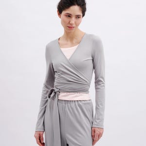 May include: A light gray long-sleeved wrap top with a V-neckline and a side tie closure, worn over a pale pink undershirt, paired with matching light gray trousers. The ensemble features a smooth, solid-colored fabric with clean lines.