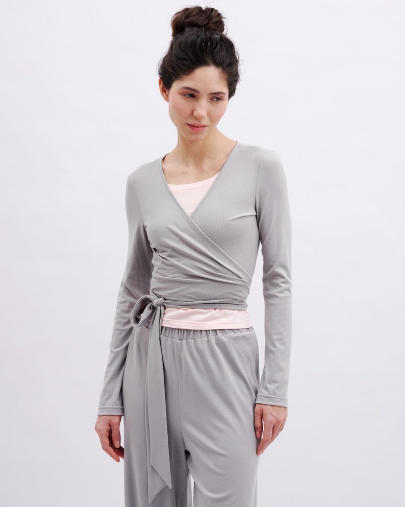 May include: A light gray long-sleeved wrap top with a V-neckline and a side tie closure, worn over a pale pink undershirt, paired with matching light gray trousers. The ensemble features a smooth, solid-colored fabric with clean lines.