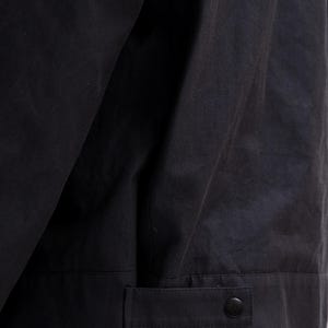 May include: Close-up of a black jacket. The jacket has a buttoned cuff detail and a button closure. The fabric appears to be a matte material. The jacket is likely part of a clothing collection.