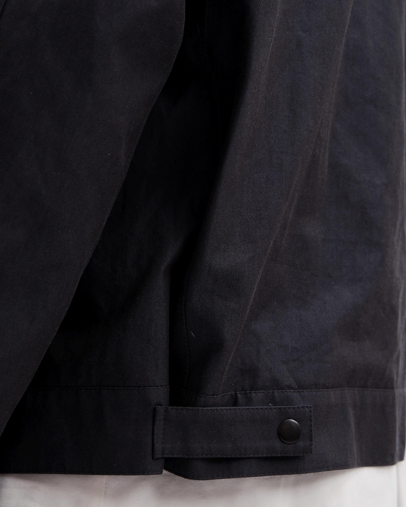 May include: Close-up of a black jacket. The jacket has a buttoned cuff detail and a button closure. The fabric appears to be a matte material. The jacket is likely part of a clothing collection.