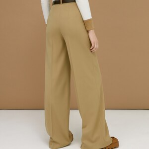 May include: Tan wide-leg trousers with a dark brown belt. The trousers have a high waist and a tailored fit. The model is wearing a white long-sleeve shirt with a brown cuff. The trousers are paired with brown shoes.