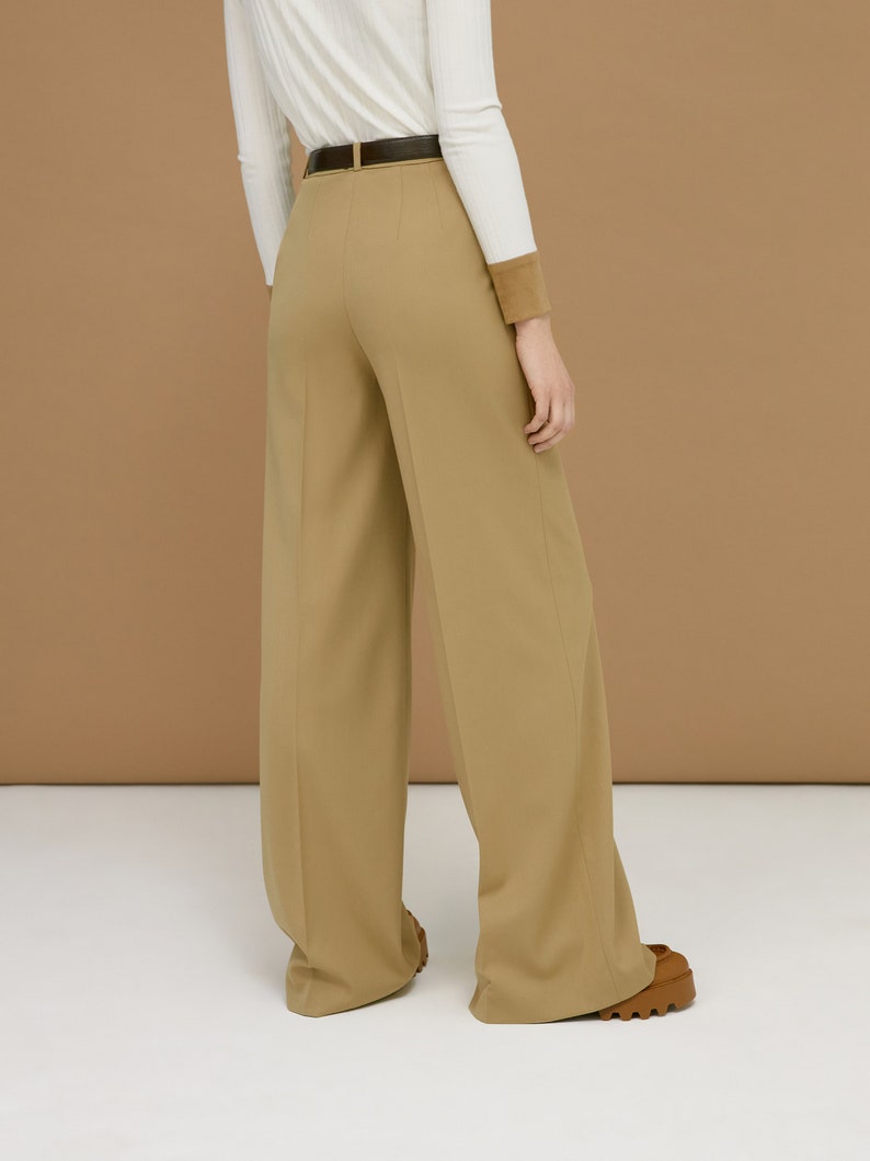 May include: Tan wide-leg trousers with a dark brown belt. The trousers have a high waist and a tailored fit. The model is wearing a white long-sleeve shirt with a brown cuff. The trousers are paired with brown shoes.