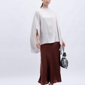 May include: A woman is wearing a light gray, loose-fitting top with a brown, flowing skirt. She is holding a small, patterned bag.