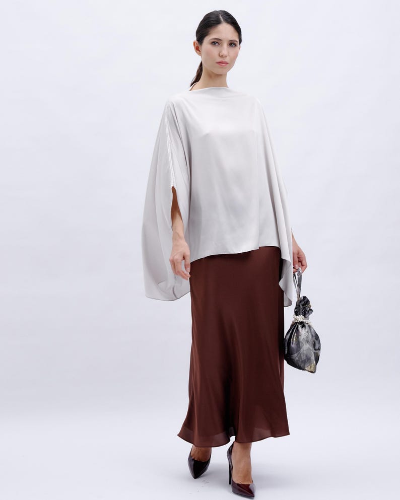May include: A woman is wearing a light gray, loose-fitting top with a brown, flowing skirt. She is holding a small, patterned bag.