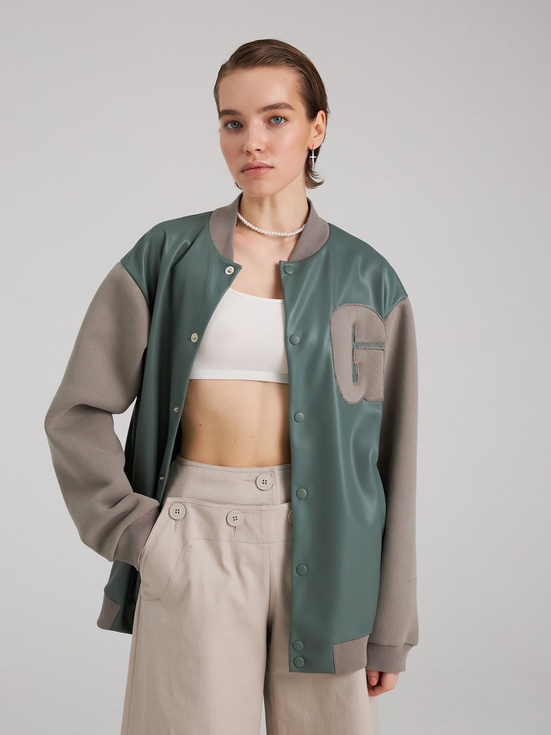 May include: A green and brown bomber jacket with a large patch on the left side. The jacket has a zipper closure and a ribbed collar.