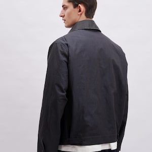 May include: A dark gray jacket with a collar and buttoned cuffs. The jacket has a simple design and a slightly shiny finish. The model is wearing a white shirt underneath and dark blue jeans.