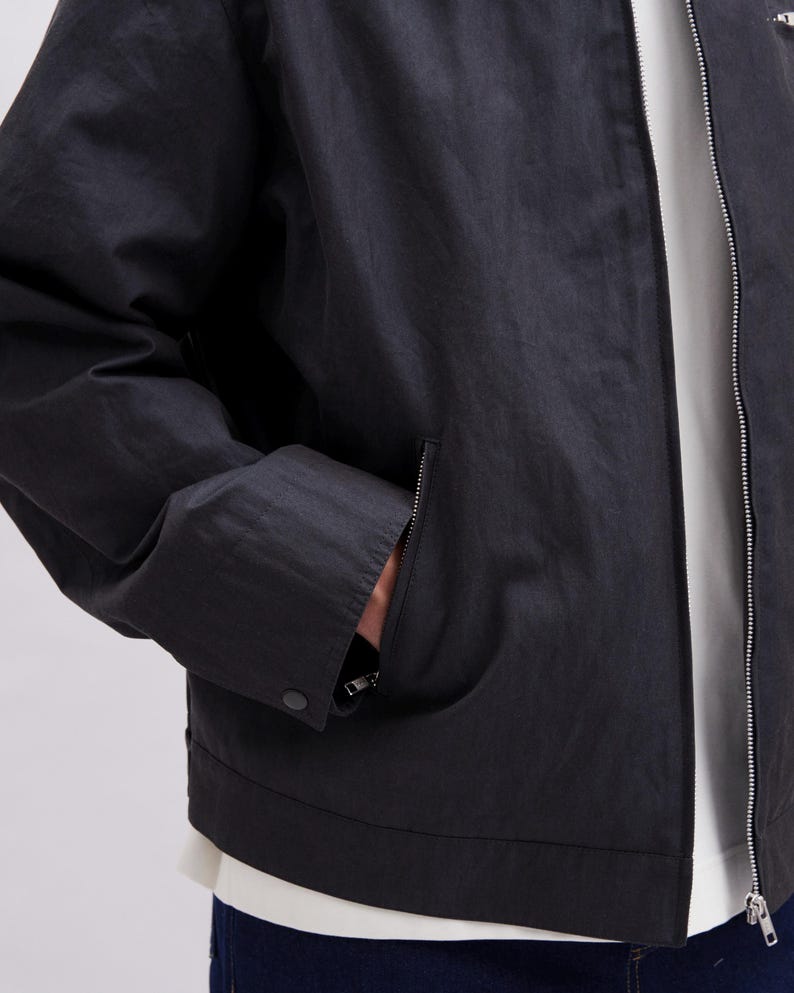 K&ouml;nnte beinhalten: Schwarze Jacke mit Rei&szlig;verschluss und einer Tasche auf der linken Seite. Die Jacke hat einen Knopfverschluss an der Tasche und einen silbernen Rei&szlig;verschluss. Die Jacke ist aus leichtem Material gefertigt.