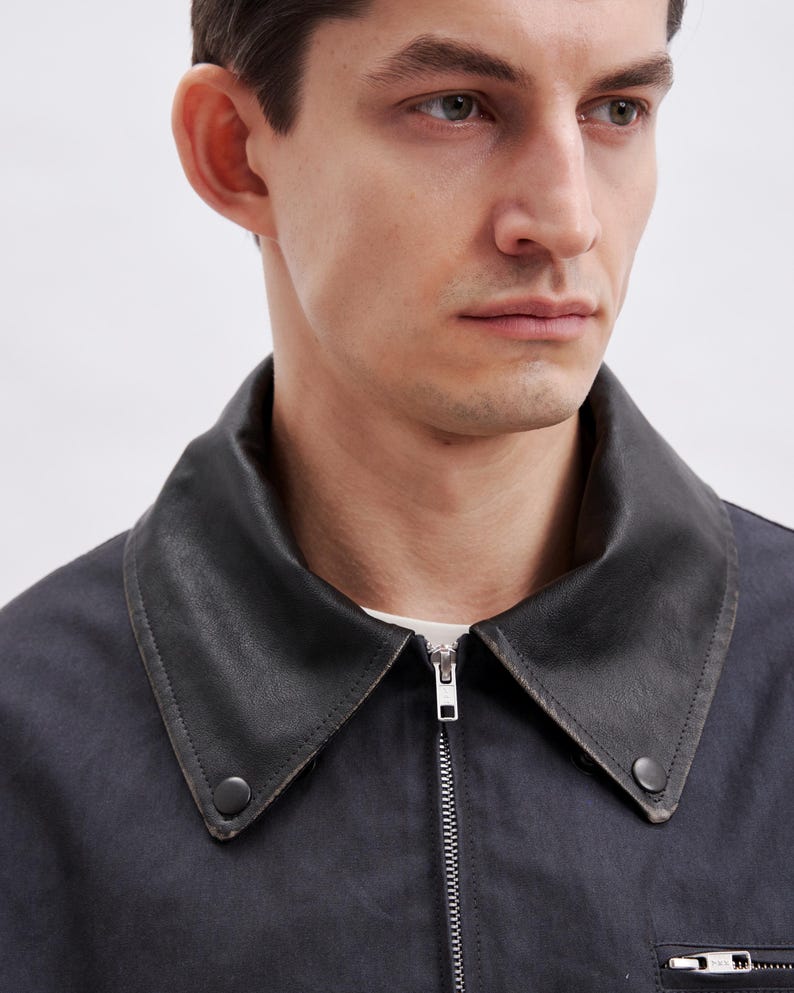 May include: A dark navy jacket with a black leather collar. The jacket has a silver zipper and a small pocket on the lower right side. The collar is buttoned on both sides.