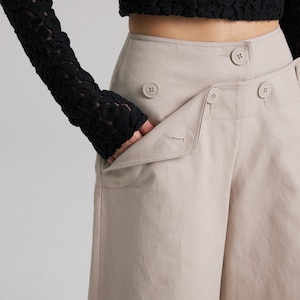 May include: Beige wide-leg pants with a high waist and multiple button closures. The pants have a relaxed fit and a slightly cropped length.