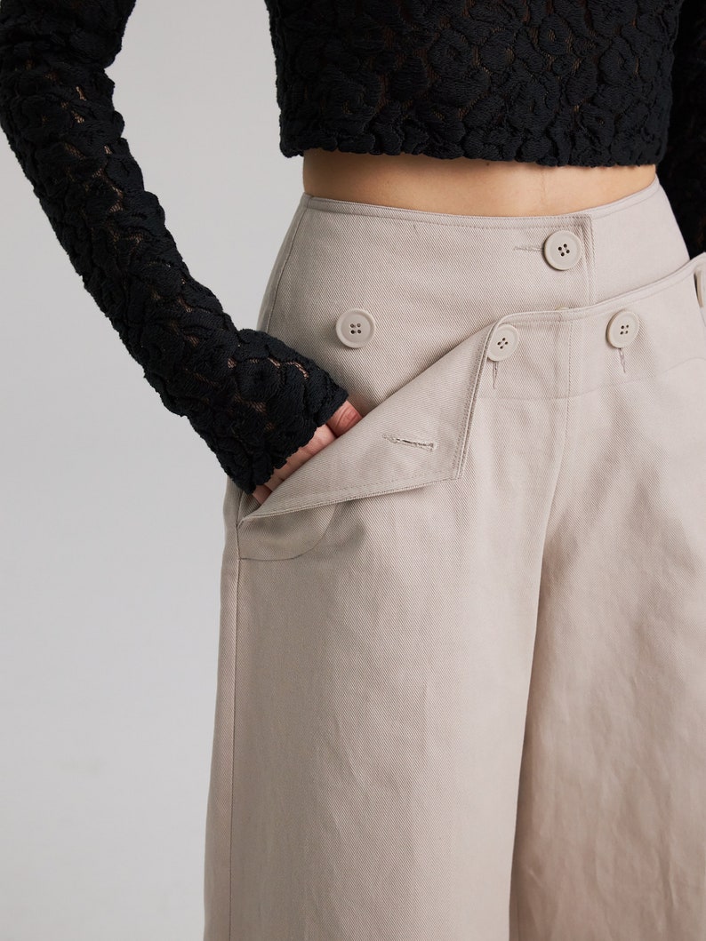 May include: Beige wide-leg pants with a high waist and multiple button closures. The pants have a relaxed fit and a slightly cropped length.