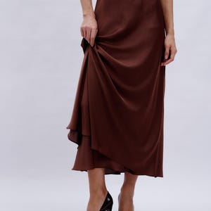 May include: A long brown satin skirt with a flowing silhouette. The skirt is worn with a white blouse and dark brown heels.