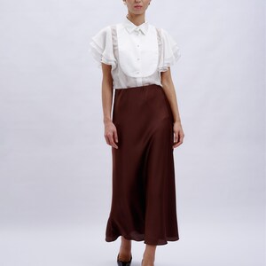 May include: A white blouse with ruffled sleeves and a brown midi skirt. The blouse has a high collar and a button-up front.