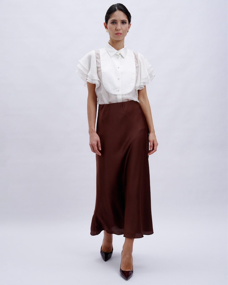 May include: A white blouse with ruffled sleeves and a brown midi skirt. The blouse has a high collar and a button-up front.