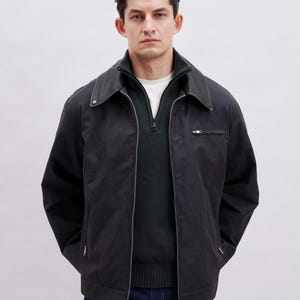 May include: A black jacket with a dark green sweater underneath. The jacket has a collar, a zipper, and a small pocket on the chest. The person is wearing a white shirt under the sweater and dark blue jeans.