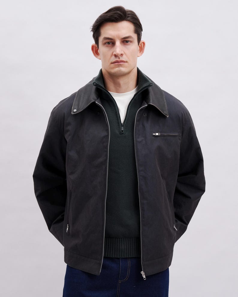 May include: A black jacket with a dark green sweater underneath. The jacket has a collar, a zipper, and a small pocket on the chest. The person is wearing a white shirt under the sweater and dark blue jeans.