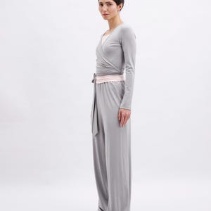 May include: A light gray long-sleeved wrap top with a matching light gray wide-leg jumpsuit featuring a pale pink waistband and tie detail. The garment is worn with pale pink ballet flats, creating a cohesive and neutral-toned ensemble.
