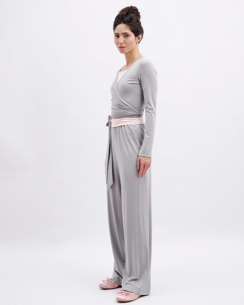 May include: A light gray long-sleeved wrap top with a matching light gray wide-leg jumpsuit featuring a pale pink waistband and tie detail. The garment is worn with pale pink ballet flats, creating a cohesive and neutral-toned ensemble.