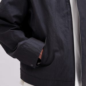 May include: A black jacket with a zipper closure and a pocket on the front. The jacket has a button closure on the pocket and a silver zipper. The jacket is made of a lightweight material.