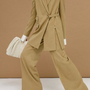 May include: A tan-colored suit with a belted blazer and wide-leg pants. The blazer has a single-button closure and a notched lapel. The outfit is paired with a white turtleneck and a cream-colored handbag. Green heels complete the look.