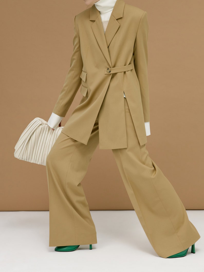 May include: A tan-colored suit with a belted blazer and wide-leg pants. The blazer has a single-button closure and a notched lapel. The outfit is paired with a white turtleneck and a cream-colored handbag. Green heels complete the look.