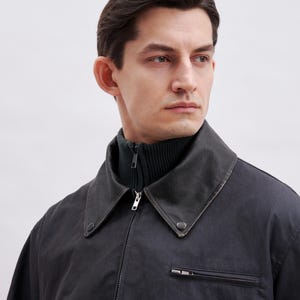 May include: A dark gray jacket with a black collar and a dark green turtleneck. The jacket has a zipper closure and a small pocket on the chest. The model is looking off to the side.