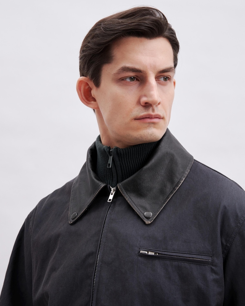 May include: A dark gray jacket with a black collar and a dark green turtleneck. The jacket has a zipper closure and a small pocket on the chest. The model is looking off to the side.