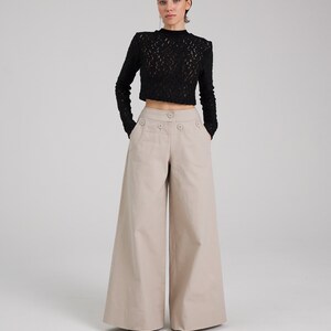 May include: A woman is wearing a black lace top and beige wide-leg pants. The pants have a high waist and a button closure.