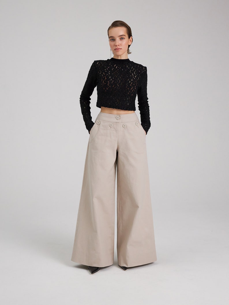 May include: A woman is wearing a black lace top and beige wide-leg pants. The pants have a high waist and a button closure.