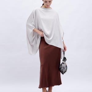 May include: A woman wearing a loose-fitting, light gray top with a brown, flowing skirt and a small, gray bag.