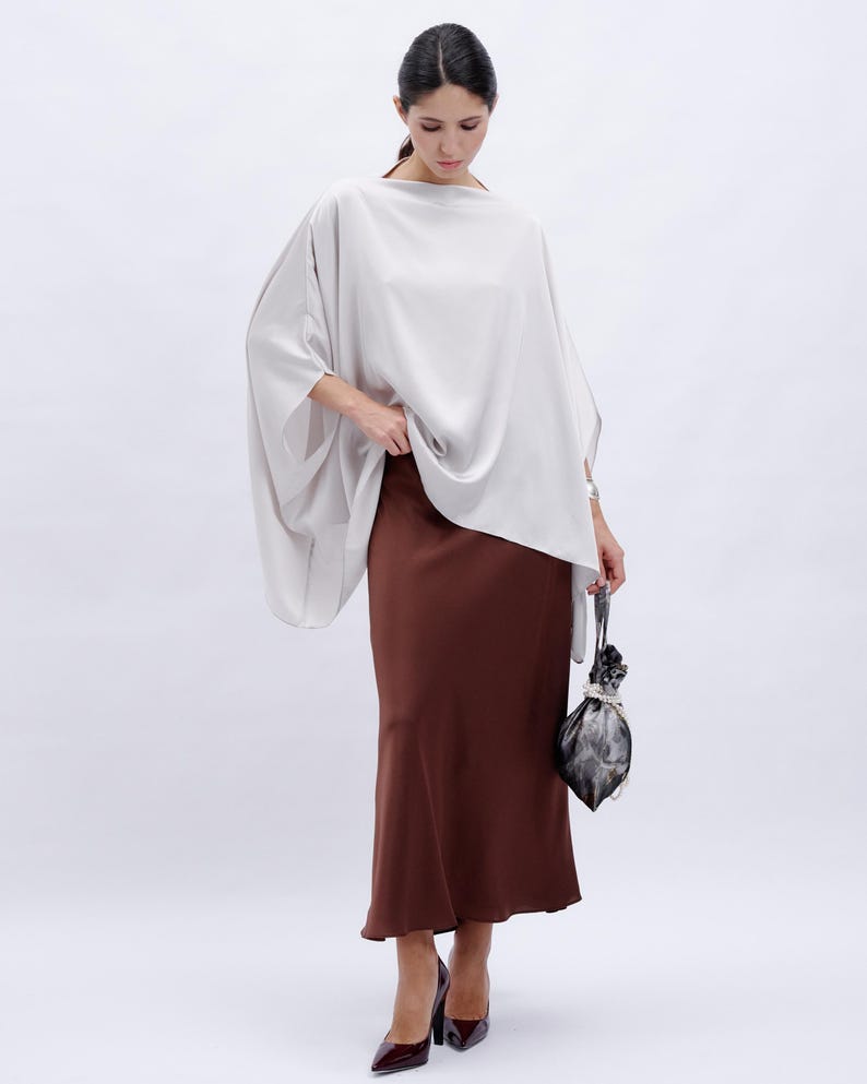 May include: A woman wearing a loose-fitting, light gray top with a brown, flowing skirt and a small, gray bag.
