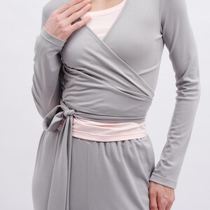 May include: A light gray long-sleeve wrap top with a side tie closure, worn over a pale pink crew neck undershirt. The wrap top features a V-neckline and fitted sleeves, paired with matching light gray trousers in a smooth, solid fabric.