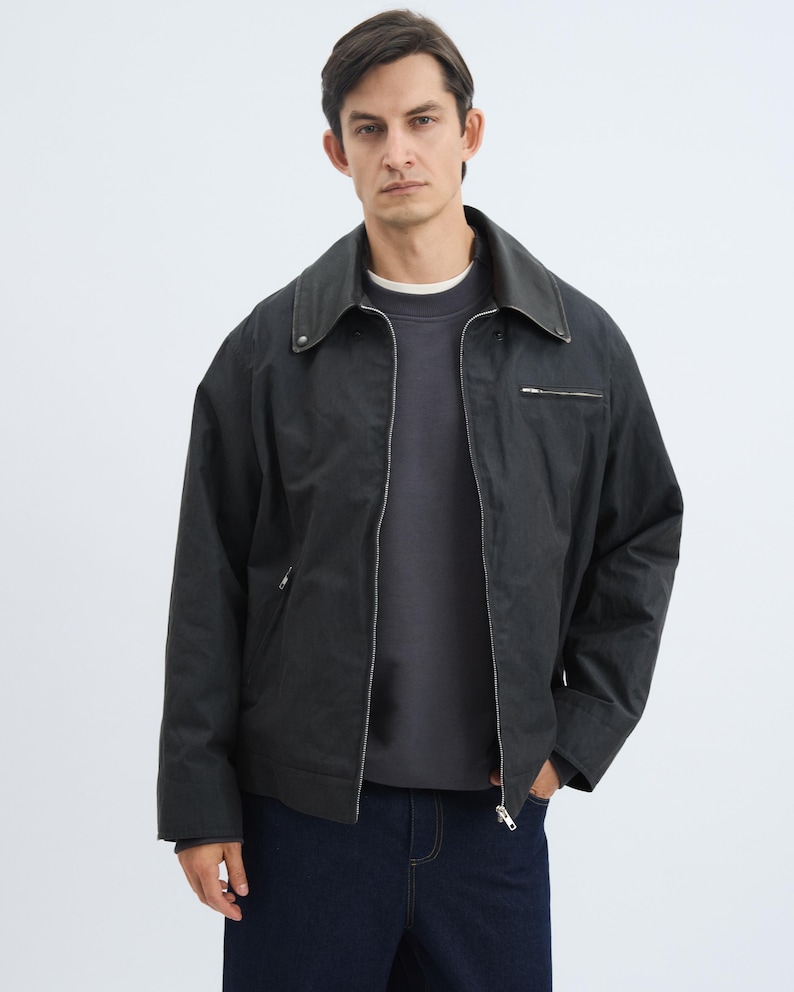 K&ouml;nnte beinhalten: Dunkelgraue Jacke mit Kragen und Rei&szlig;verschluss. Die Jacke wird &uuml;ber einem dunkelgrauen Sweatshirt getragen und mit dunkelblauen Jeans kombiniert. Das Model steht vor einem wei&szlig;en Hintergrund.