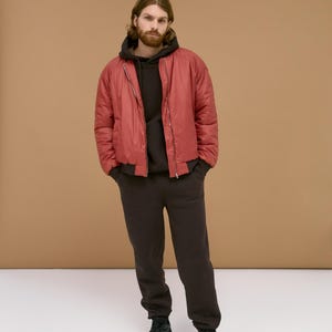 May include: A red bomber jacket with a front zipper, worn over a dark brown hooded sweatshirt and matching dark brown sweatpants. The outfit is completed with black athletic sneakers, featuring a solid color design and a casual, relaxed fit.