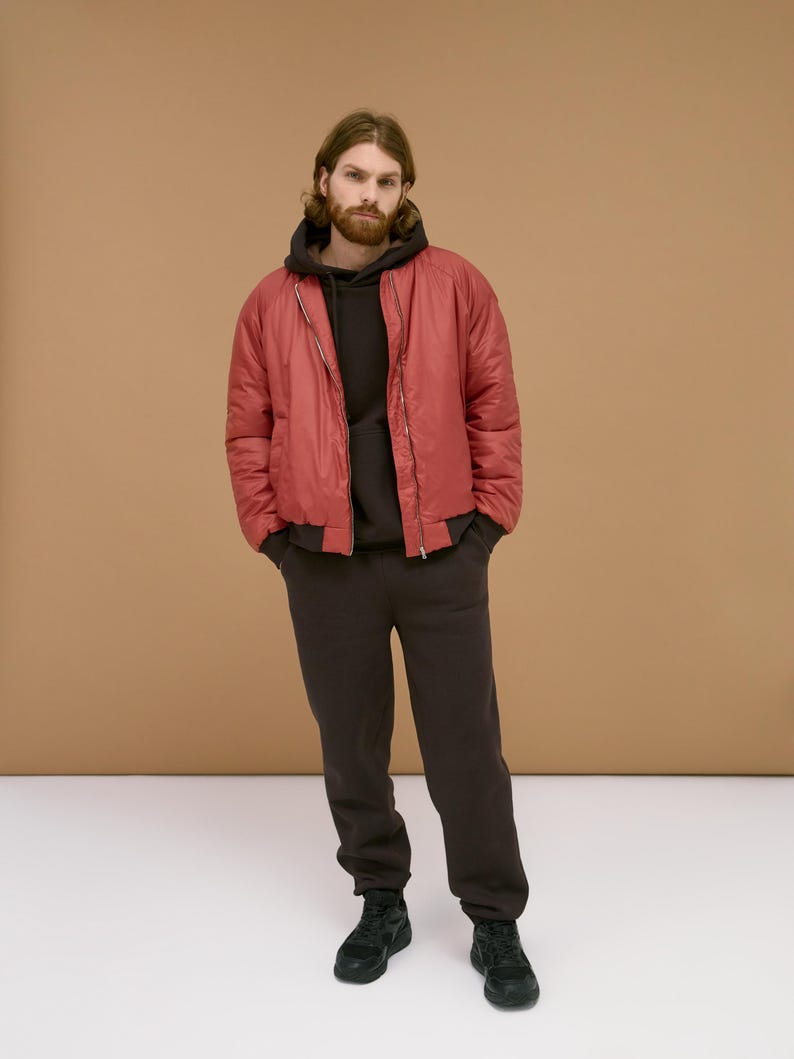 May include: A red bomber jacket with a front zipper, worn over a dark brown hooded sweatshirt and matching dark brown sweatpants. The outfit is completed with black athletic sneakers, featuring a solid color design and a casual, relaxed fit.