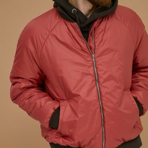 May include: A red bomber jacket with a smooth finish, featuring a silver front zipper, long sleeves, and dark brown ribbed cuffs and hem. The jacket is worn over a dark brown hooded sweatshirt with a visible drawstring and metal eyelets.