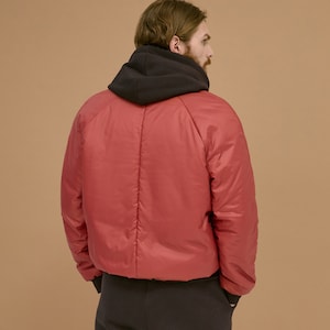 May include: A red puffer jacket with long sleeves and a black hood layered underneath. The jacket features a smooth, quilted texture with a central vertical seam on the back, paired with black trousers that have a simple, clean design.