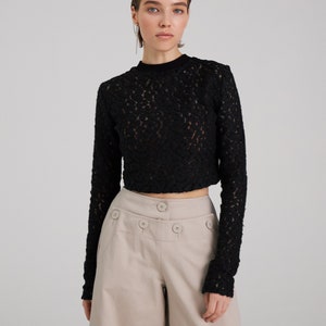 May include: A black lace long-sleeve top with a high neckline and a cropped hem. The top is paired with beige high-waisted pants with multiple buttons.