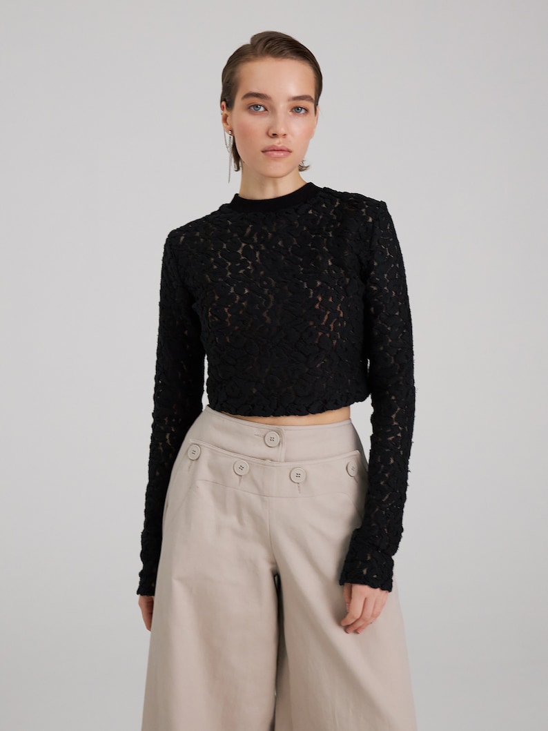 May include: A black lace long-sleeve top with a high neckline and a cropped hem. The top is paired with beige high-waisted pants with multiple buttons.