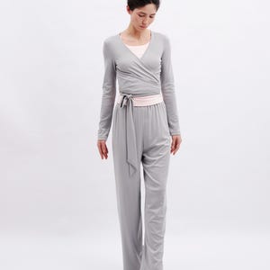 May include: A light gray long-sleeved wrap top with a tie waist and matching wide-leg trousers. The outfit is layered over a pale pink scoop-neck undershirt. The ensemble is paired with soft, pale pink ballet-style slippers with a small bow.