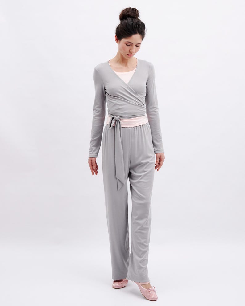 May include: A light gray long-sleeved wrap top with a tie waist and matching wide-leg trousers. The outfit is layered over a pale pink scoop-neck undershirt. The ensemble is paired with soft, pale pink ballet-style slippers with a small bow.