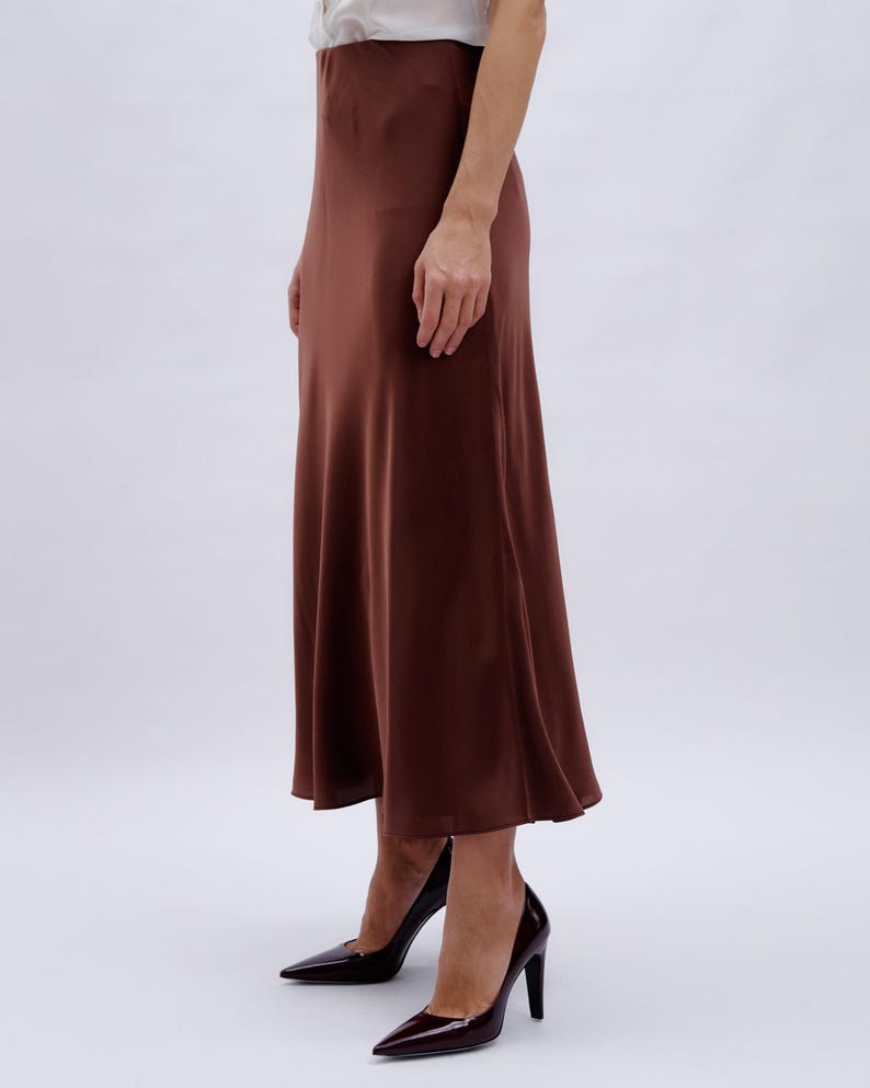 May include: A brown satin midi skirt with a slight A-line shape. The skirt is worn with a pair of dark burgundy heels.