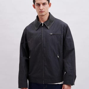 May include: A dark gray jacket with a zip-up front and a contrasting dark collar. The jacket has two side pockets and a small chest pocket. The model is wearing a white shirt underneath and dark blue jeans.