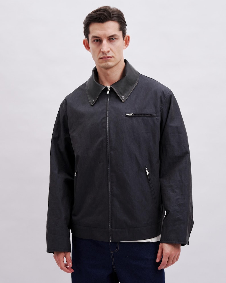 May include: A dark gray jacket with a zip-up front and a contrasting dark collar. The jacket has two side pockets and a small chest pocket. The model is wearing a white shirt underneath and dark blue jeans.